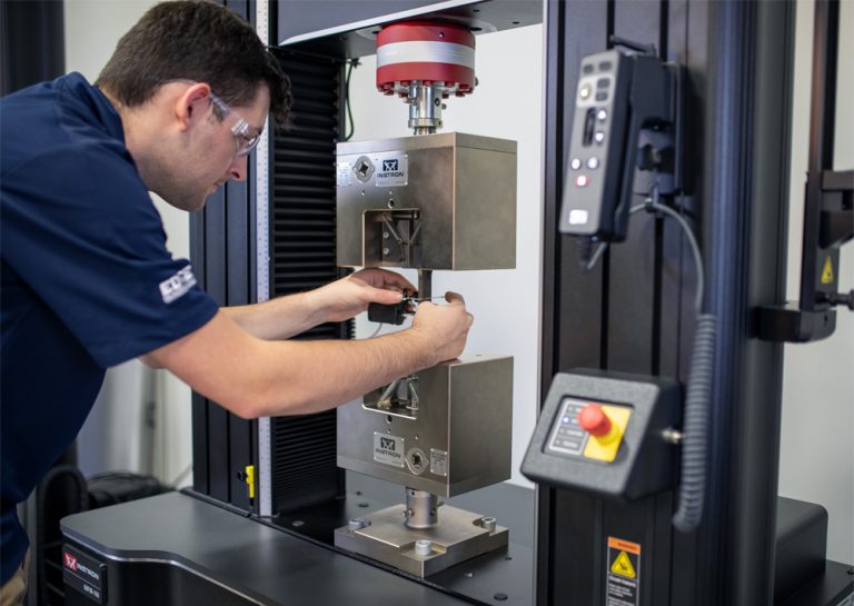 Operator in safety glasses carefully attaching a clip-on extensometer to a metal tensile specimen clamped in Instron wedge grips on a 68FM-100 universal testing machine