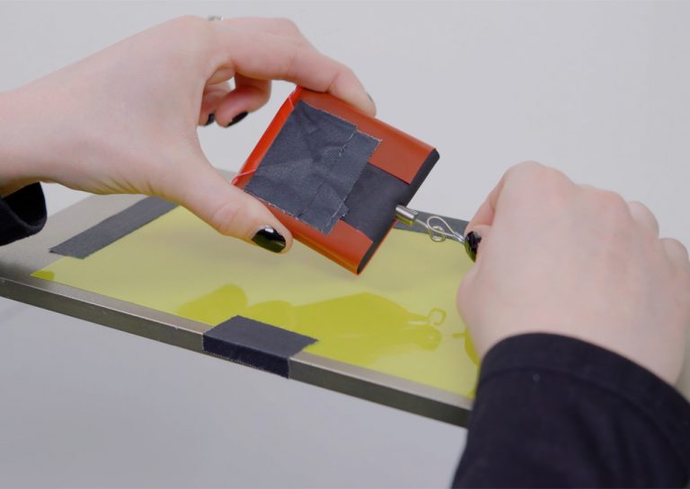 Close-up of a lab operator's hands connecting a tow line hook to a weighted sled wrapped with a film specimen on a yellow film specimen taped to the horizontal test platform of an Instron coefficient of friction fixture