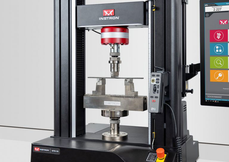 Three-point flexure fixture with a metal test specimen positioned on an Instron 68TM-100 table model universal testing system, with the Bluehill Universal software operator dashboard in view.
